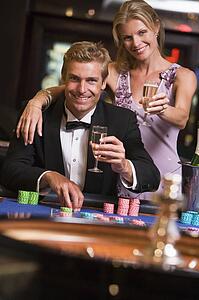 Couple in casino at roulette table holding up champagne and smiling (selective focus)