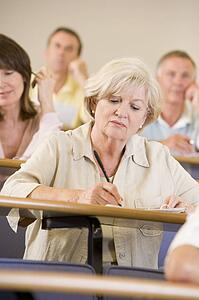 Woman sitting in adult classroom taking notes with students in background (selective focus)
