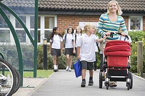 Woman and young boy pushing a stroller outside school with students in background (selective focus)
