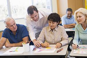 Adult students in class drawing pictures with teacher helping (selective focus)