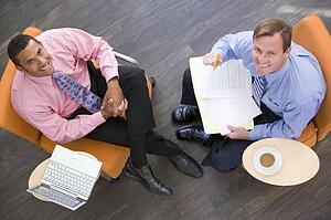 Two businessmen sitting indoors with coffee laptop and folder smiling