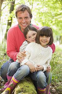 Father and daughters outdoors in woods sitting on log smiling