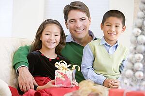 Father And His Son And Daughter Sitting On Sofa, Holding A Christmas Present