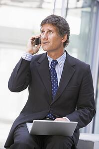 Businessman sitting in office lobby with laptop using cellular phone