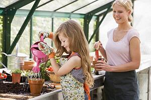 Young girl in greenhouse watering plant with woman holding pot smiling