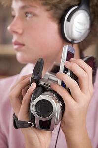 Young boy wearing headphones in bedroom holding many electronic devices