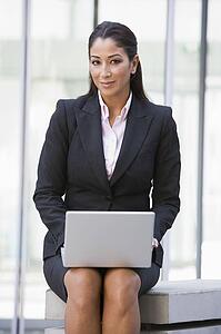 Businesswoman sitting outdoors by building with laptop smiling (high key/selective focus)