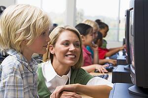 Teacher helping student at computer terminal with students in background (depth of field/high key)