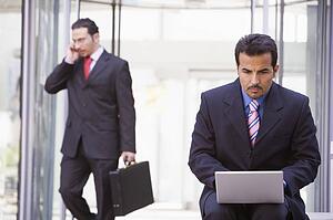 Businessman outdoors in front of building using laptop with businessman in background on cellular phone (high key/selective focus)