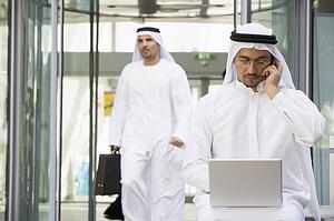Businessman outdoors with laptop on cellular phone with businessman walking in background (selective focus)
