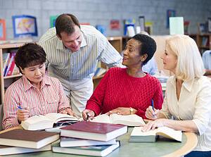 Three women sitting in library with books and notepads while a man leans over them (selective focus)