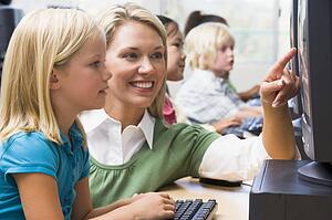 Teacher helping student at computer terminal with students in background (depth of field/high key)