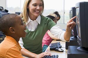 Teacher helping student at computer terminal with students in background (selective focus/high key)