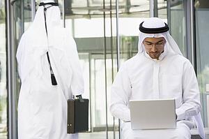 Businessman outdoors using laptop with businessman walking in background (selective focus)