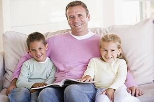 Man and two children sitting in living room reading book and smiling