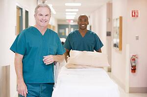 Two Orderlies Pushing An Empty Bed Down A Hospital Corridor