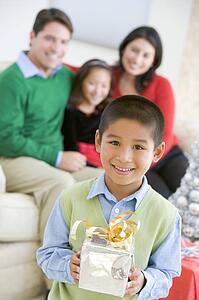 Young Boy Standing Holding Christmas Present, With His Parents And Sister In The Background