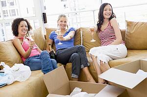 Three girl friends relaxing with champagne by boxes in new home smiling