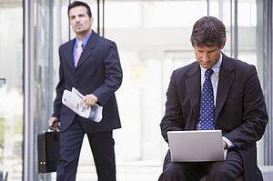 Businessman outdoors in front of building using laptop with businessman in background carrying newspaper (high key/selective focus)