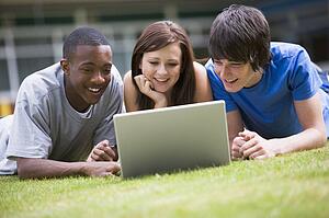 Three students lying outdoors on lawn with laptop