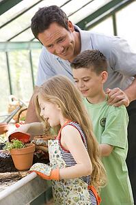 Man in greenhouse helping two young children putting soil in pot