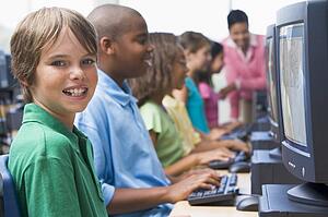 Six children at computer terminals with teacher in background (depth of field/high key)