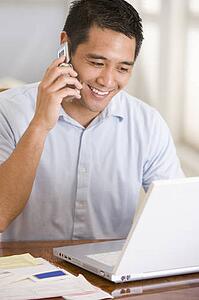 Man in dining room on cellular phone using laptop smiling