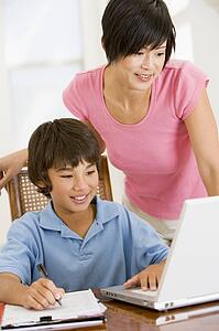 Woman helping young boy with laptop do homework in dining room smiling