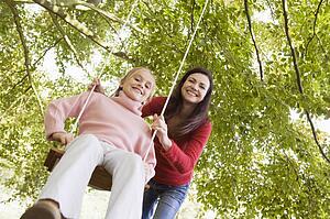 Woman outdoors pushing young girl on swing and smiling (selective focus)