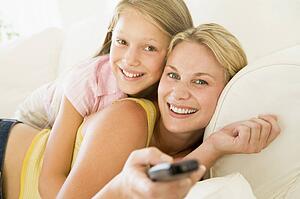 Woman and young girl with remote control embracing on sofa smiling