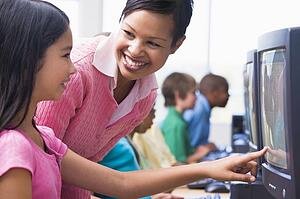 Teacher helping student at computer terminal with students in background (depth of field/high key)