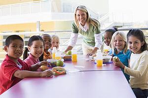 Teacher leaning on table outdoors while students eat lunch (high key)
