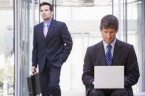 Businessman outdoors in front of building using laptop with businessman in background (high key/selective focus)