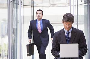 Businessman outdoors in front of building using laptop with businessman running in background (high key/selective focus)