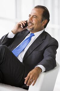 Businessman sitting indoors on cellular phone smiling (high key/selective focus)