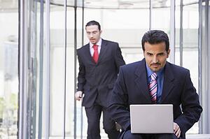 Businessman outdoors in front of building using laptop with businessman in background (high key/selective focus)