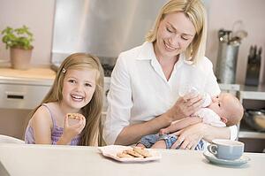 Mother feeding baby in kitchen with daughter eating cookies and smiling
