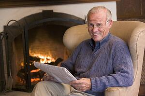 Man sitting in living room by fireplace with newspaper smiling