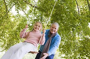 Grandfather outdoors pushing granddaughter on swing and smiling (selective focus)