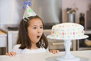 Young girl wearing party hat at kitchen counter looking at cake smiling