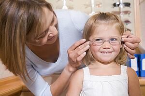 Woman trying eyeglasses on young girl at optometrists smiling