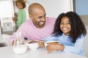 Father Sitting With Daughter As She They Eat Breakfast With Her Mother In The Background 