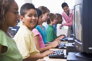 Six children at computer terminals with teacher in background (selective focus/high key)