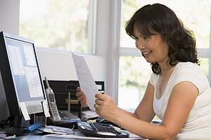 Woman in home office with computer and paperwork smiling