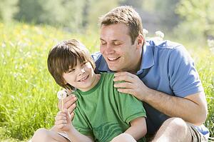 Father and son sitting outdoors with dandelion head smiling