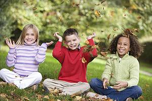 Three young children sitting outdoors in park throwing leaves in air and smiling (selective focus)
