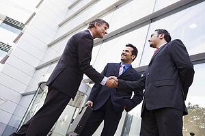 Three businessmen standing outdoors by building shaking hands and smiling (high key/selective focus)