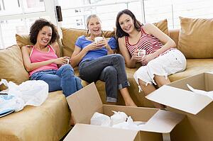 Three girl friends relaxing with coffee by boxes in new home smiling