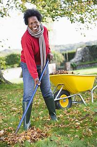 Woman outdoors raking leaves near wheelbarrow and smiling (selective focus)