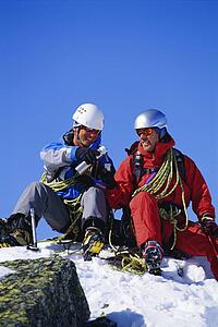Two mountain climbers sitting on snowy mountain drinking from thermos and smiling (selective focus)